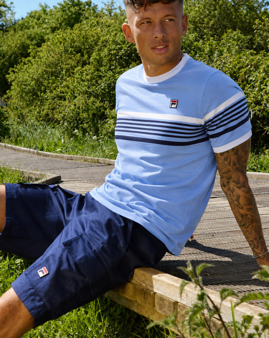 Male model relaxing on a wooden boardwalk in a sunny outdoor setting, wearing a light blue FILA striped T-shirt and navy Fila shorts.