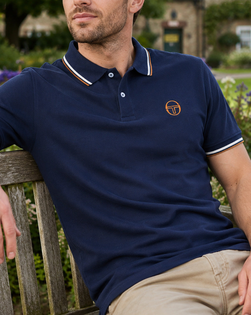 Male model wearing a navy Sergio Tacchini tipped polo shirt, relaxing on a wooden bench in an English garden.
